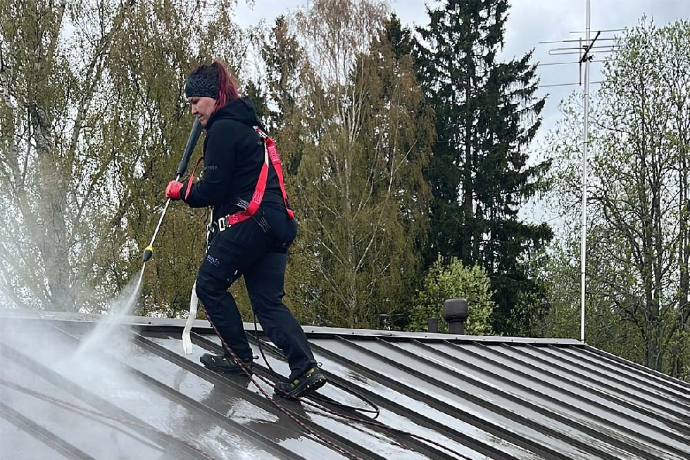 Henkilö pesee peltikattoa painepesurilla valjaiden turvin.